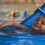 Niño feliz disfrutando sus clases de natación con un flotador azul, promoviendo seguridad en el agua y desarrollo temprano en la niñez